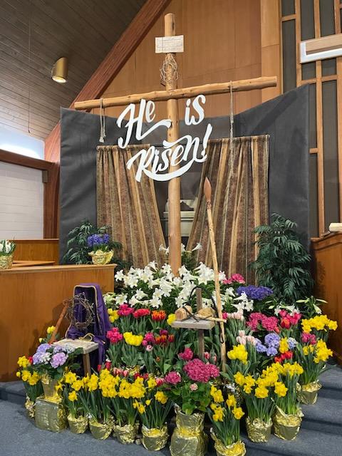 A display of Easter celebration with "He is Risen" written on the cross and flowers around the base of the cross.